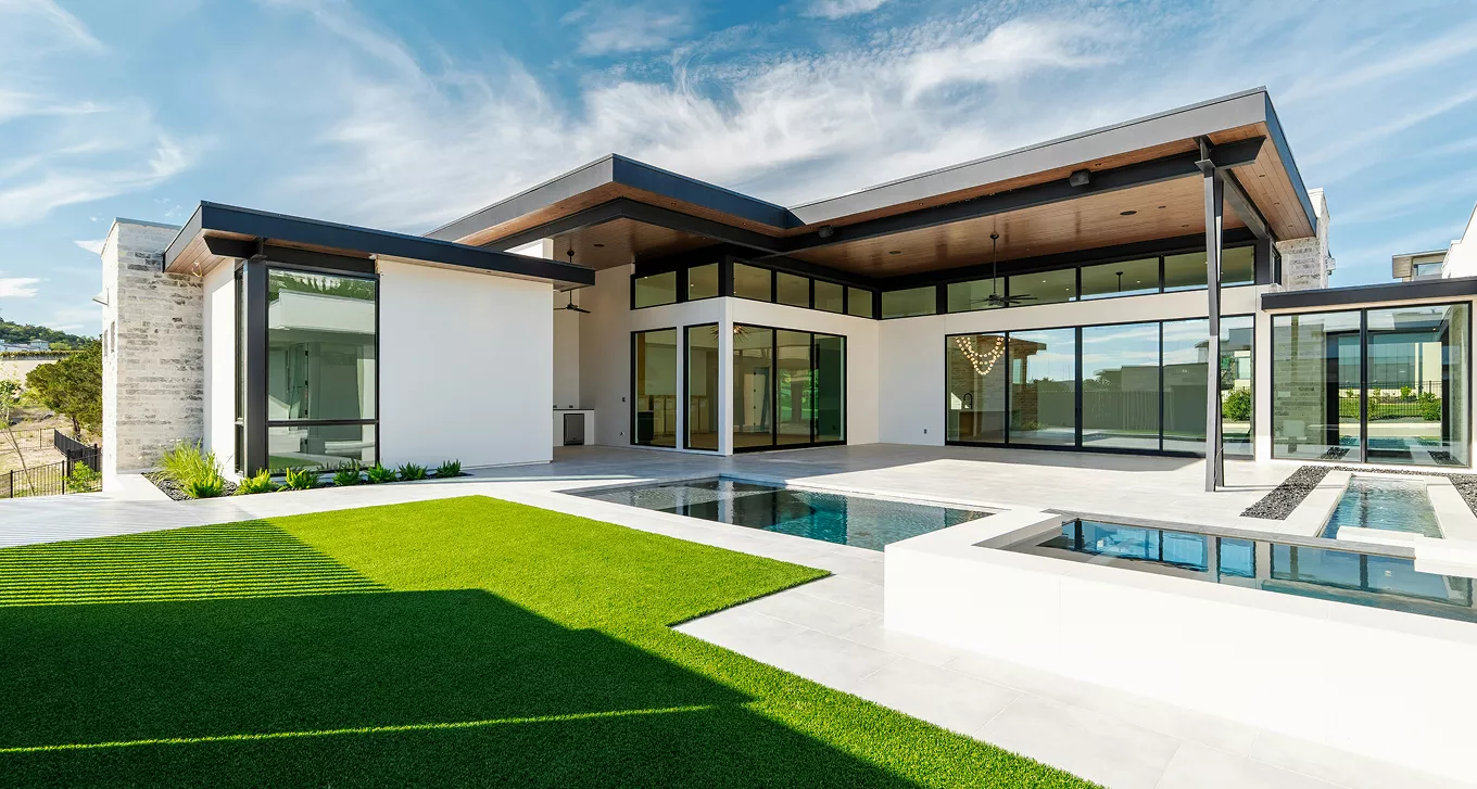 Modern flat-roof home with wraparound glass walls, wood ceiling accents, and a sleek backyard infinity pool.
