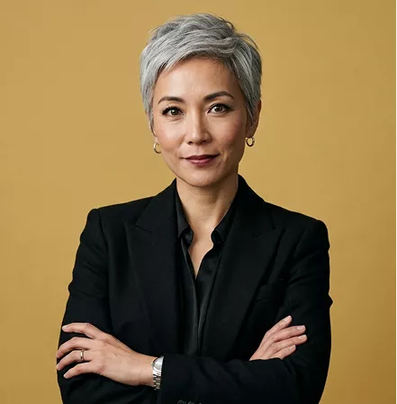 Professional headshot of a woman with short gray hair and a black blazer, arms crossed, on a mustard background.