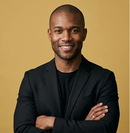 Professional headshot of a smiling bald man with a black blazer, arms crossed, on a mustard background.