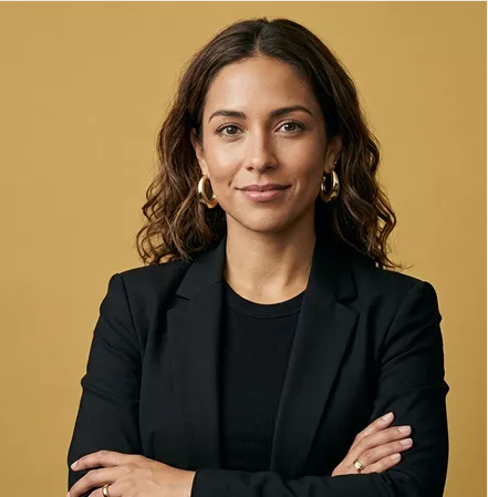 Professional headshot of a woman with wavy brown hair and gold hoop earrings, wearing a black blazer, on a mustard background.