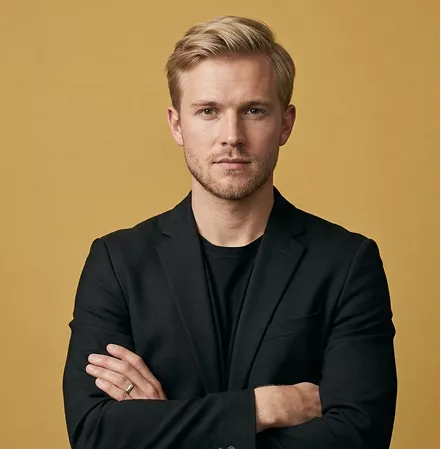 Professional headshot of a man with short blond hair and stubble, wearing a black blazer, arms crossed, on a mustard background