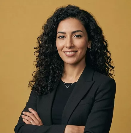 Professional headshot of a smiling woman with curly dark hair and a black blazer, arms crossed, on a mustard background.